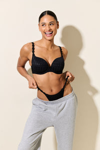 Woman wearing a black bra and underwear set with gray sweatpants on a beige background